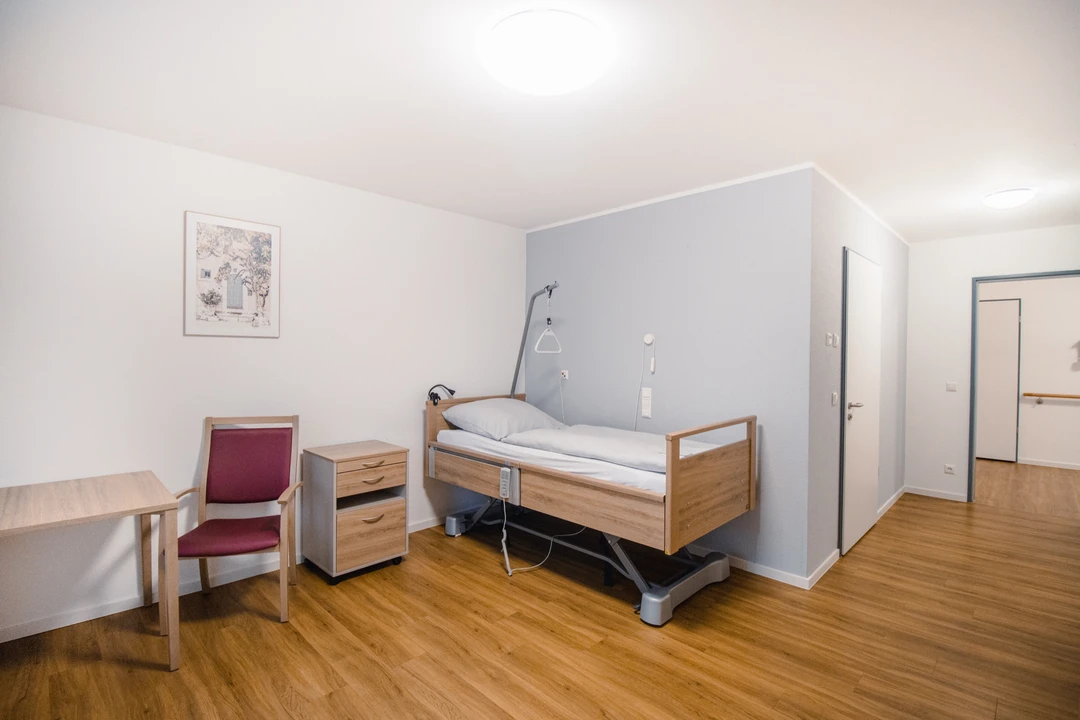Einzelzimmer mit höhenverstellbarem Pflegebett, Sessel, Nachttisch und Schreibtisch
